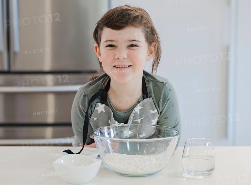 A cheerful child wearing an apron smiles while preparing to bake in a modern kitchen. The setup includes clear bowls with flour and water, showcasing a delightful cooking scene