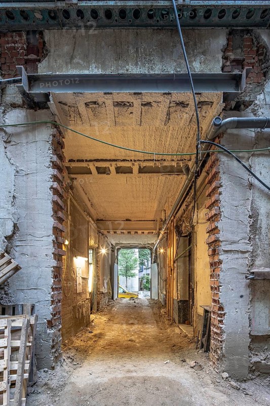A construction site featuring a partially demolished entrance with exposed bricks and reinforced beams. The unfinished passageway leads to an open courtyard, highlighting ongoing restoration efforts