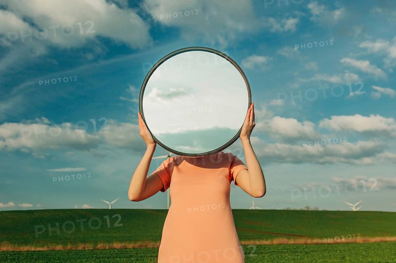 A person stands in a field, holding a circular mirror that reflects the blue sky and clouds, creating a surreal and thought provoking visual effect with nature