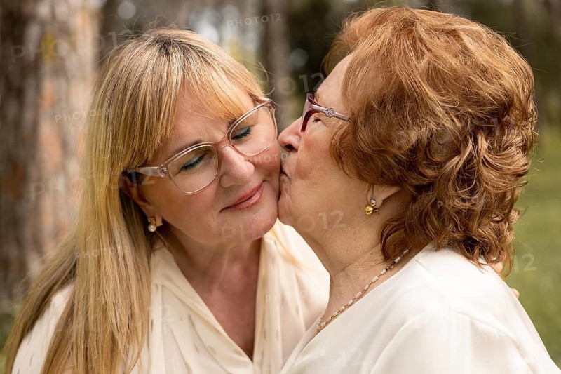 An elderly mother lovingly kisses her adult daughter on the cheek, showcasing the warmth and affection inherent in their relationship. The backdrop is a serene outdoor setting