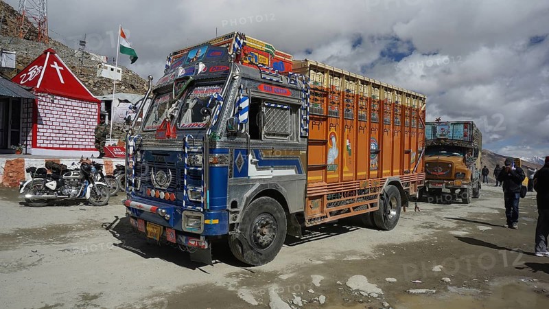 Colourful truck at Khardung-La Pass next to other vehicles and small houses with Indian flag under cloudy sky, trekking in Ladakh, Himalayas, India