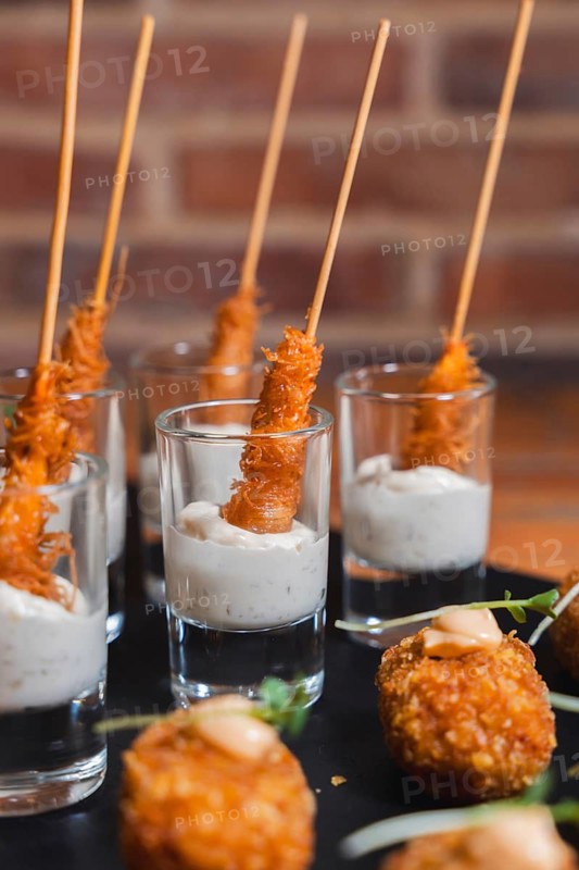 Close-up of crispy skewered appetizers artfully served in shot glasses filled with a creamy dipping sauce, elegantly arranged on a black slate platter against a rustic brick wall