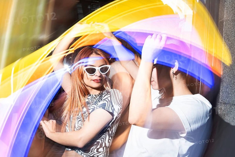 A vibrant, artistic photo of a lesbian couple enjoying a colorful light display. They are smiling, with bright, swirling lights enhancing the lively atmosphere