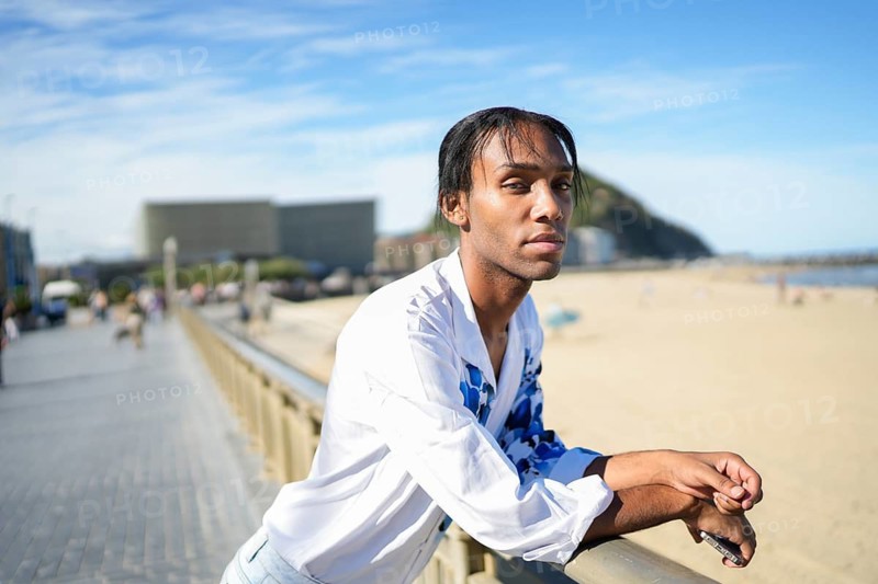 A man wearing a white shirt stands by a beach railing, confidently expressing LGBTQIA+ pride. The sunny setting adds a warm, welcoming atmosphere to the scene
