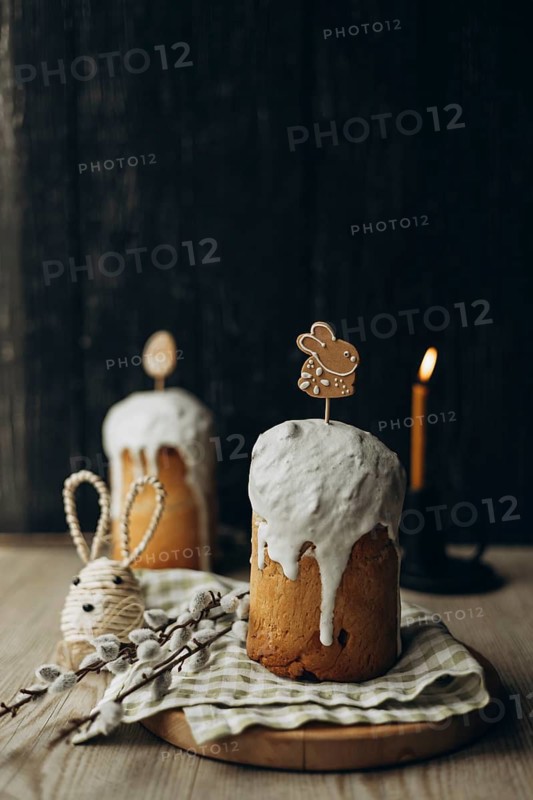 Kulich, a traditional Russian Easter bread, adorned with icing and playful gingerbread cookie toppers, sits on a rustic table accompanied by candlelight, willow catkin branches and Easter themed decorations