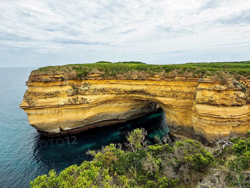 A stunning coastal rock formation forming a natural arch over azure waters, surrounded by lush green vegetation in the Twelve Apostles, Australia. The cloudy sky adds depth to this serene landscape