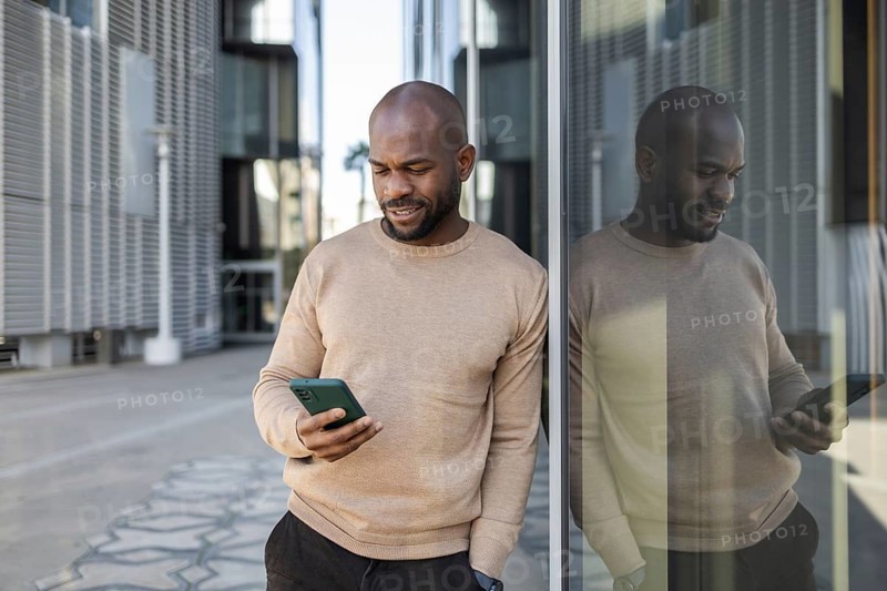 A cuban man in a beige sweater leans against a glass building facade, engrossed in his smartphone. The reflection creates a symmetrical visual effect, capturing a serene moment in an urban setting