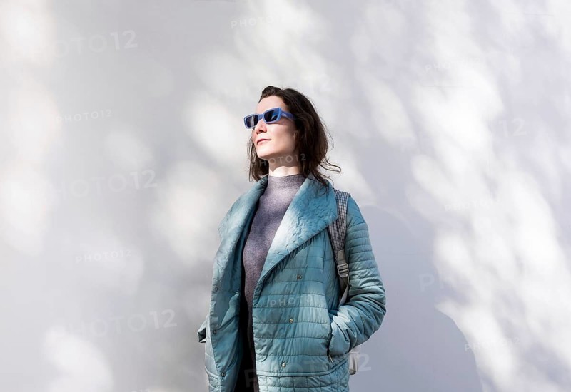 A woman in a light blue jacket and sunglasses stands against a white wall, bathed in sunlight and shadows of leaves, embodying the spirit of spring