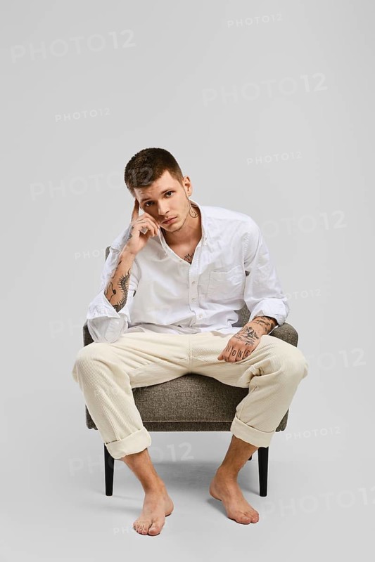Full length studio portrait of young barefoot man in white shirt and ivory trousers sitting in armchair