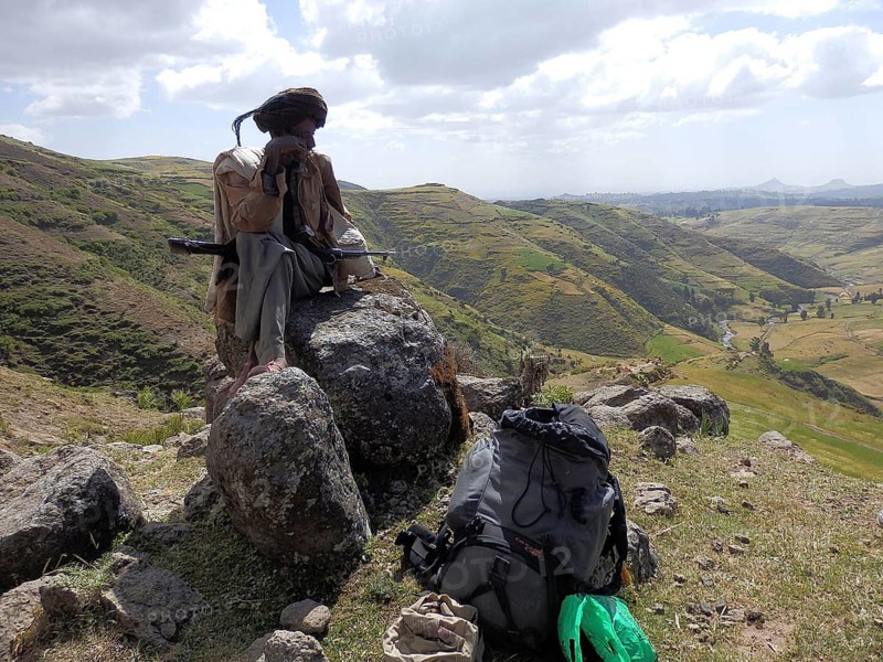 Man sitting with his rifle, Kalashnikov, on a rock with backpack in the foreground, surrounded by mountainous landscape, Simien National Park, Ethiopia