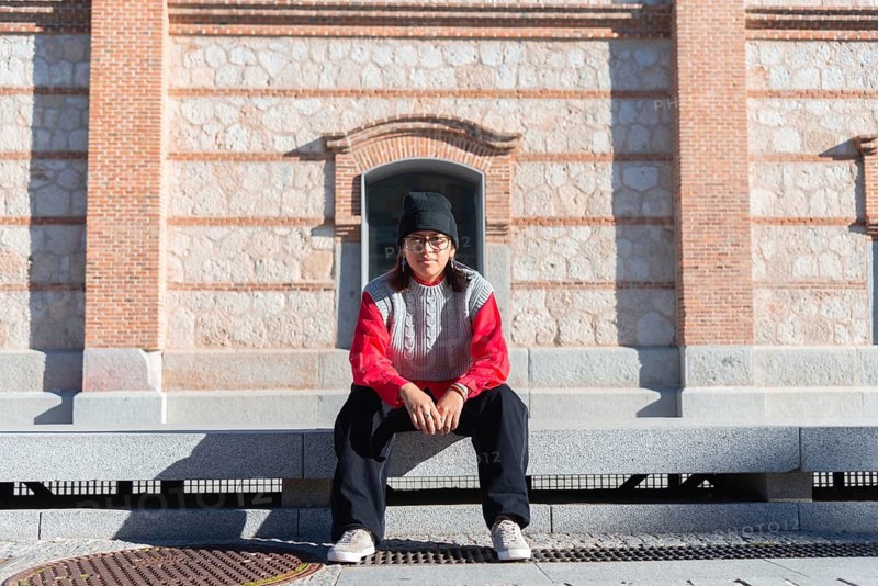 A latin young adult in casual streetwear sits on an urban bench With a modern style, the scene captures a blend of contemporary fashion and urban architecture