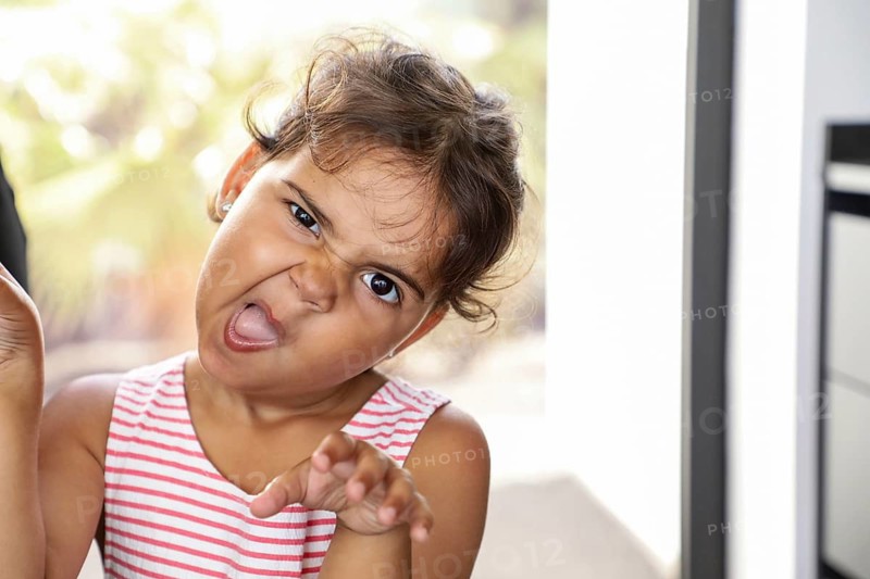 A playful child in a striped top strikes a silly face and gestures dramatically while indoors. The natural light enhances the vibrant expression, capturing youthful joy