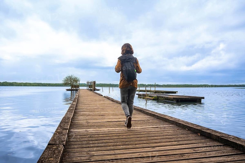 Woman wearing a backpack walking down a rustic wooden pier stretching into lake azur under a cloudy sky, finding adventure and solitude while traveling in france