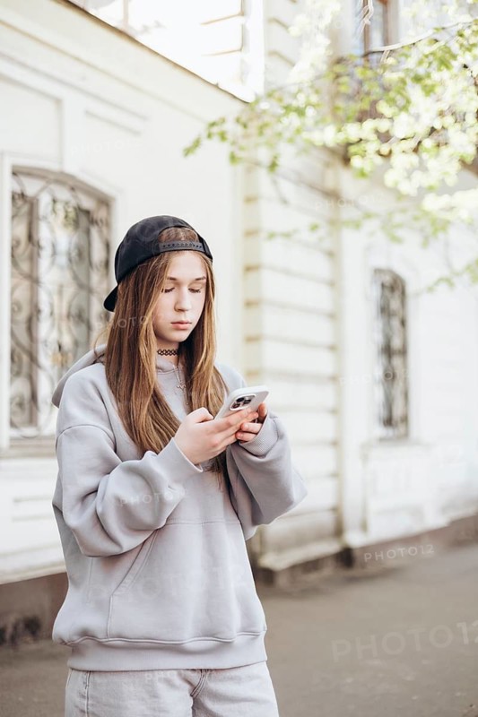 Teen girl in casual outfit with a black cap focuses intently on her smartphone. The outdoor setting is relaxed, with daylight highlighting her concentration on the device