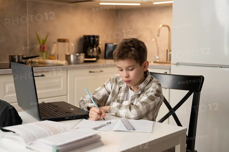 A young boy concentrates on his homework at a kitchen table. Surrounding him are a laptop and notebooks, symbolizing technology and learning in a cozy, modern kitchen setting