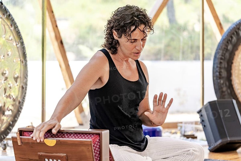 A female sound therapist with eyes closed plays a Shruti Box during a peaceful sound therapy session. The serene setting includes surrounding gongs, enhancing the holistic ambiance