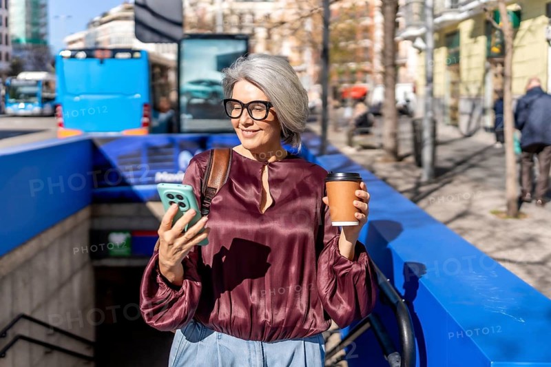 Woman standing on a busy city street near a subway entrance, observing content on her mobile phone while holding a disposable coffee cup, reflecting urban lifestyle and technology