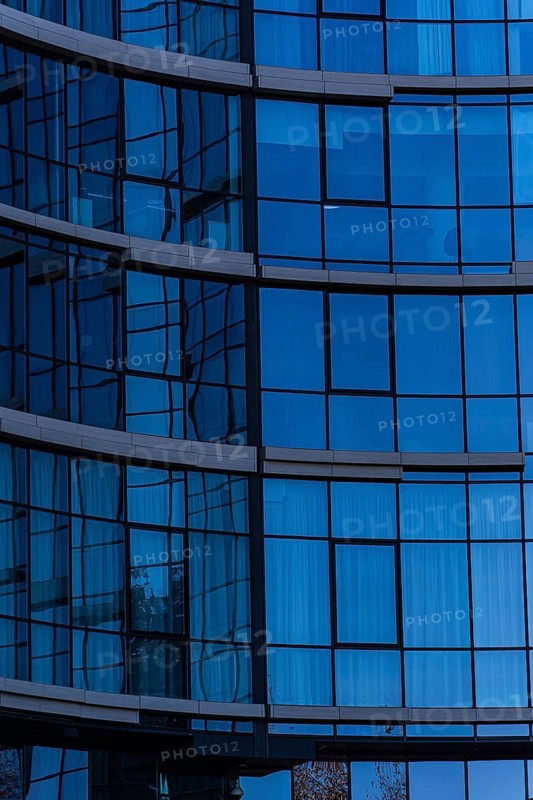 A close-up view of a modern office building, showcasing a curved glass facade with visible metal supports and reflections. The architecture uses concrete, glass, and metal elements for a sleek and contemporary look