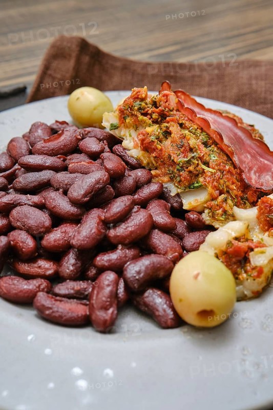 A plate features a hearty portion of red beans paired with stuffed meat that includes herbs, vegetables, and slices of cured meat. Green olives add a touch of acidity to the meal