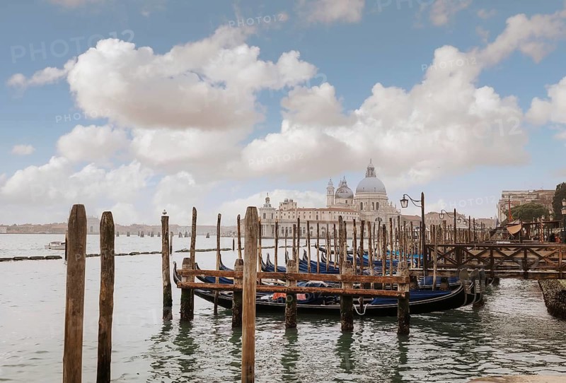 Gondolas are moored along wooden piers in Venice, Italy. The iconic Basilica di Santa Maria della Salute is visible in the background under a vibrant, partly cloudy sky