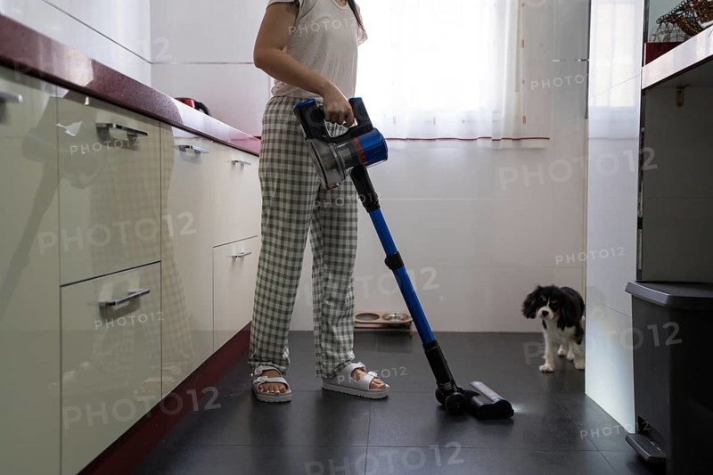 Woman uses a cordless vacuum cleaner in a bright kitchen, maintaining cleanliness and hygiene. A small dog curiously observes the activity from a corner of the room