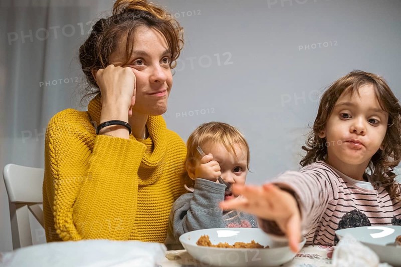 A mother, looking tired yet content, sits at a dining table with her two young children during breakfast time. The scene captures warmth, family bonding, and daily routines