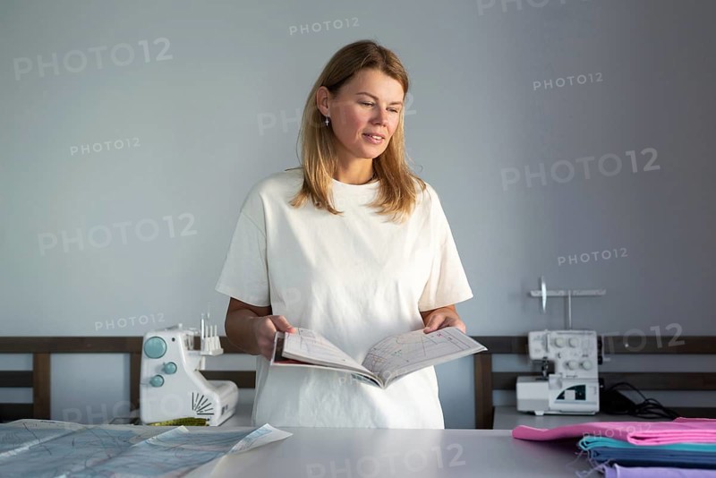 Woman in a bright sewing workshop, reviewing a pattern guide with sewing machines and colorful fabrics. The workspace reflects creativity and meticulous craftsmanship