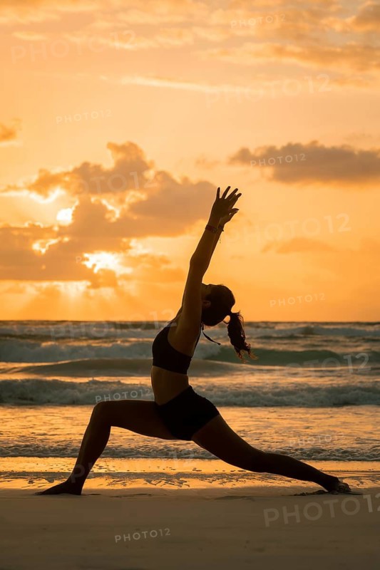A silhouette of a woman performing yoga on the beach as the sun sets, casting a warm glow in Tulum, Mexico. The calming waves and colorful sky create a serene and peaceful scene