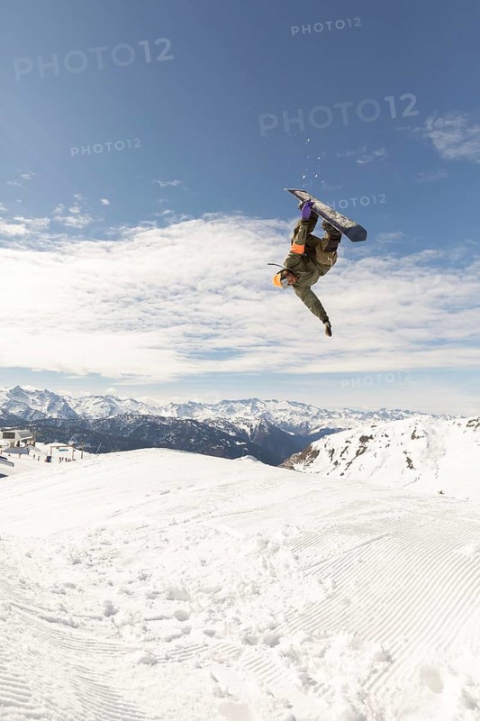 Dynamic photo of a snowboarder performing an aerial trick against a backdrop of snowy mountains. The action captures the thrill of snowboarding and winter sports adventure