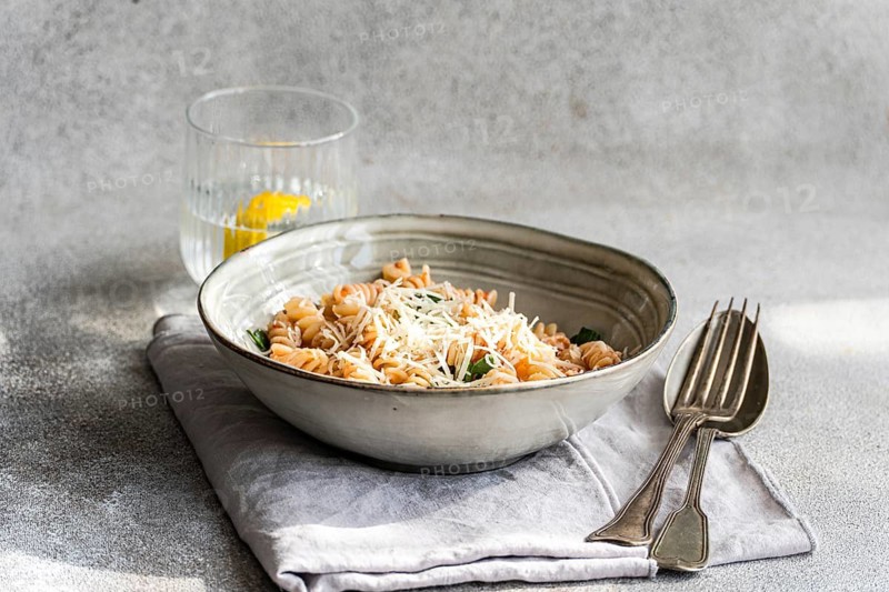 A ceramic bowl of cavatappi pasta topped with cheese and spicy tomato sauce, served on a napkin alongside cutlery, and a refreshing lemon water on a textured surface
