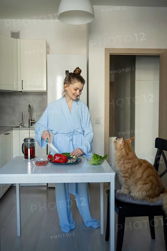 A woman in a blue robe smiles while chopping vegetables in a modern kitchen. Her orange cat attentively watches from a chair, creating a cozy domestic scene