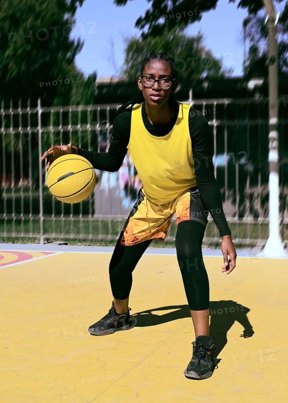 A woman athlete in a yellow vest practices dribbling on an outdoor basketball court. The scene highlights determination and focus, with vibrant colors and dynamic movement