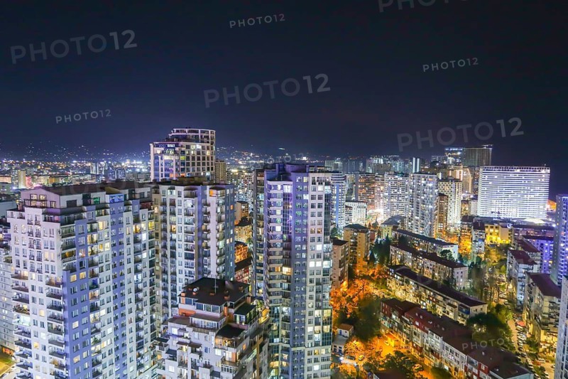 A stunning nighttime view of Batumi, Georgia, showcasing illuminated high-rise buildings and vibrant streets. The skyline is set against a backdrop of distant hills under a starry sky