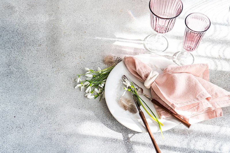 Top view of a stylish table setting featuring fresh snowdrops, a white ceramic plate, pink cloth napkins, and elegant pink glassware on a speckled surface