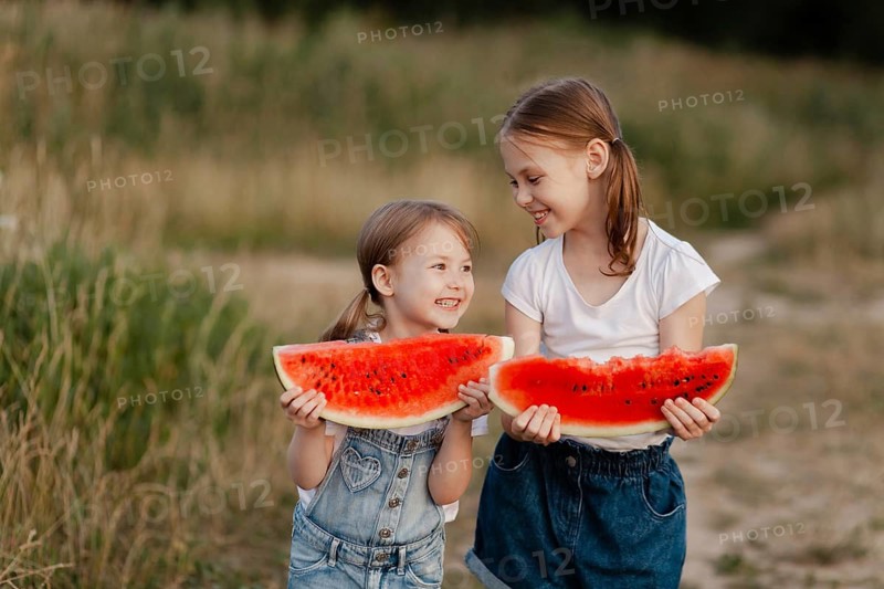 Two smiling children enjoy large watermelon slices outdoors in a sunlit field. Dressed in casual summer attire, they laugh and share a joyful moment of refreshment and play