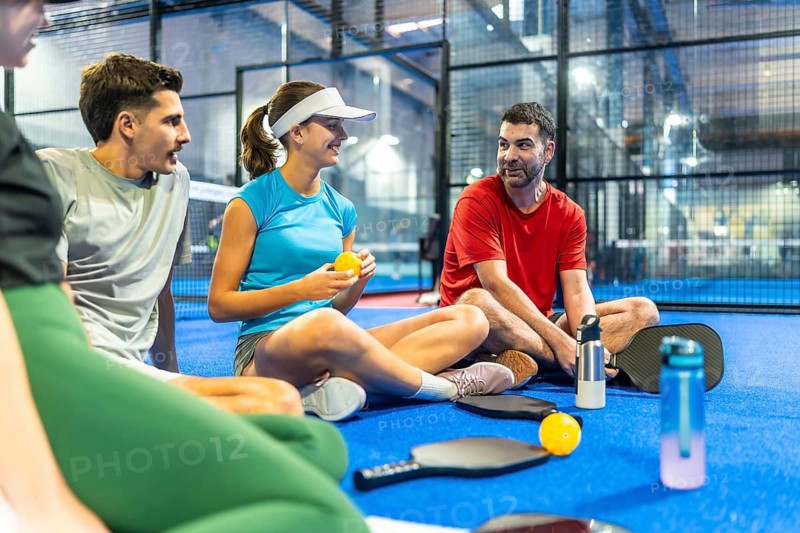 Group of diverse friends relaxing and chatting together on the blue pickleball court surface, holding paddles and balls, enjoying leisure time and camaraderie