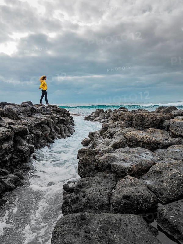 A woman in a yellow jacket stands on rocky terrain by the ocean at Timanfaya National Park, Lanzarote. Dramatic sky and waves enhance the rugged coastal landscape