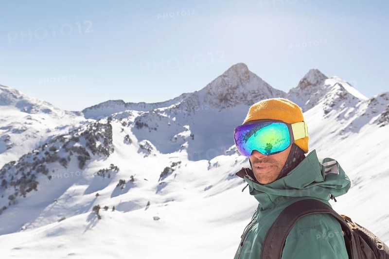 A snowboarder dressed in winter gear stands confidently in a backcountry mountain setting. The sunlight enhances the snowy landscape, ideal for a freeride adventure