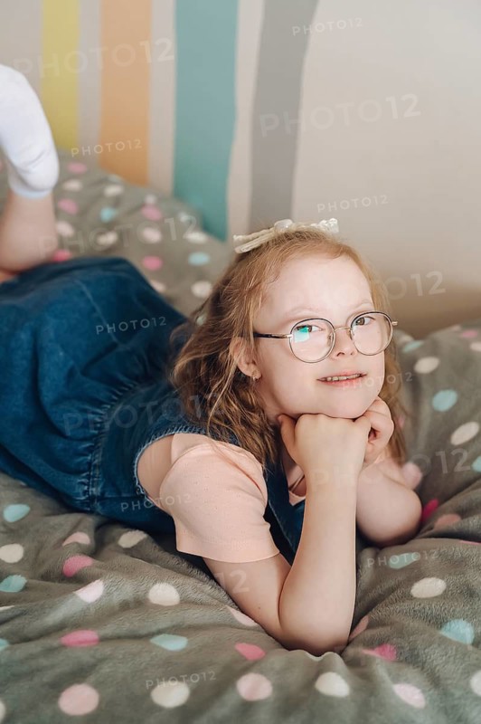 A young girl with Down syndrome joyfully engages in playtime in a colorful room. She smiles, wearing glasses and a denim dress, embodying happiness and warmth