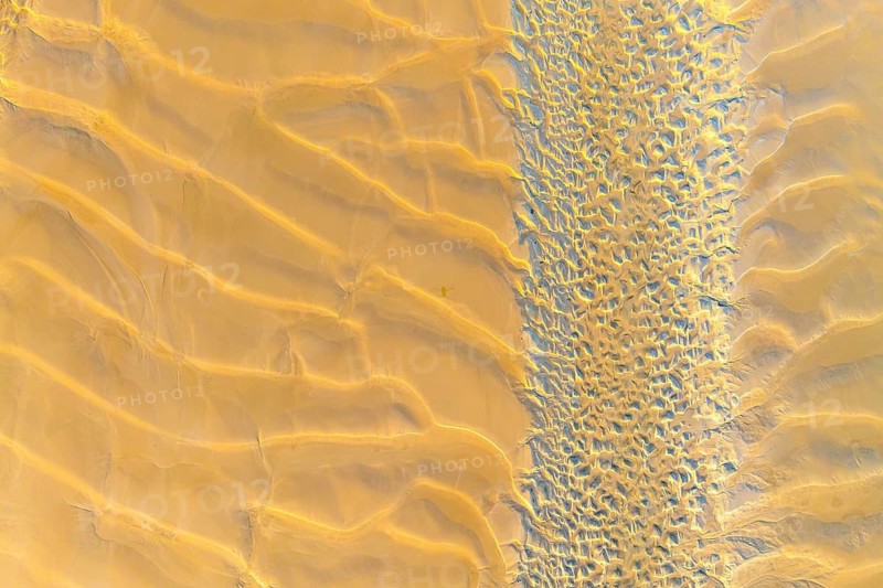 An aerial photograph showcases vast, golden sand dunes and intricate textures in a desert landscape. Shadows and sunlight create a stunning, natural abstract design