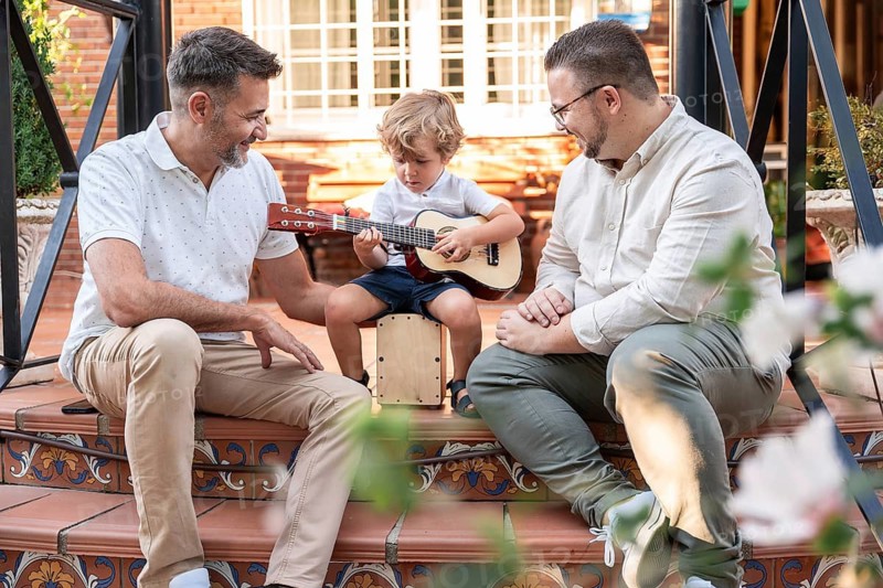 A joyful gay couple shares a musical moment with their young son, who is playing a small guitar. They sit on decorative steps, fostering love and family bonding outdoors