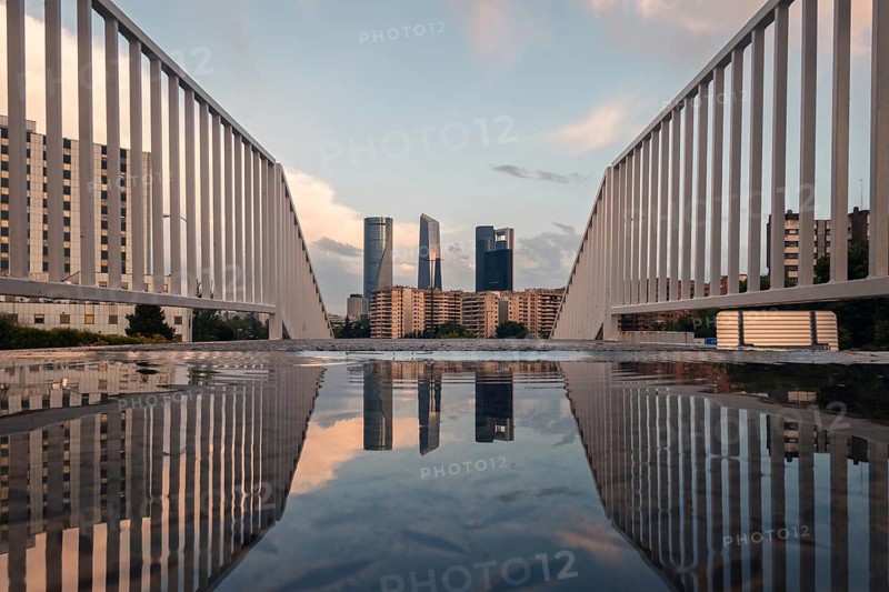 Dramatic view from a modern bridge in Madrid capturing the city skyline reflected in a puddle at sunset. The sky is filled with scattered clouds creating a stunning scene