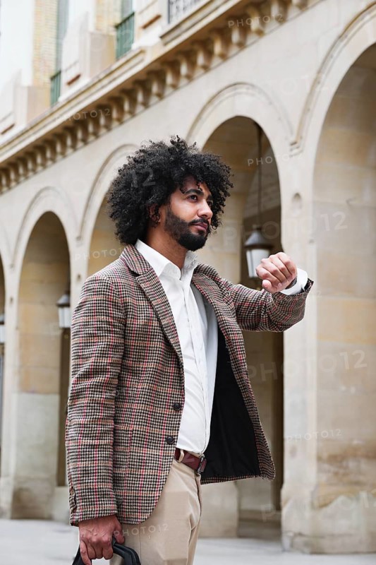 Mixed-race insurance salesman with afro hair, wearing a plaid blazer and white shirt, checks his watch while standing in front of a stone building with arches