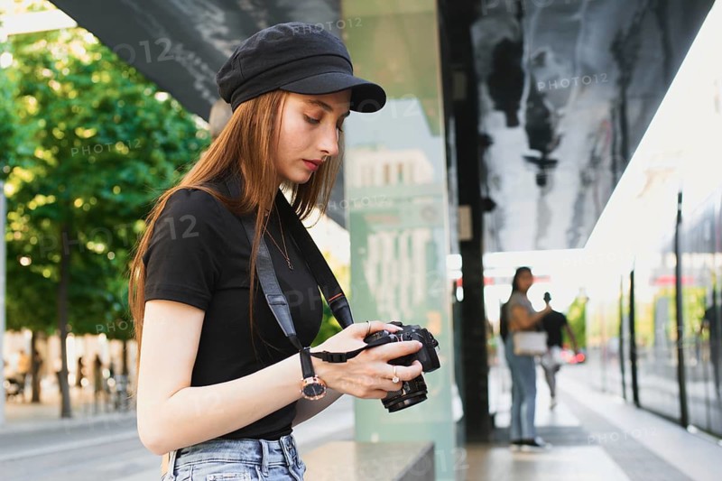 A young woman photographer focuses intently on her camera, capturing moments in a vibrant city setting. She wears casual attire, blending seamlessly with the urban landscape