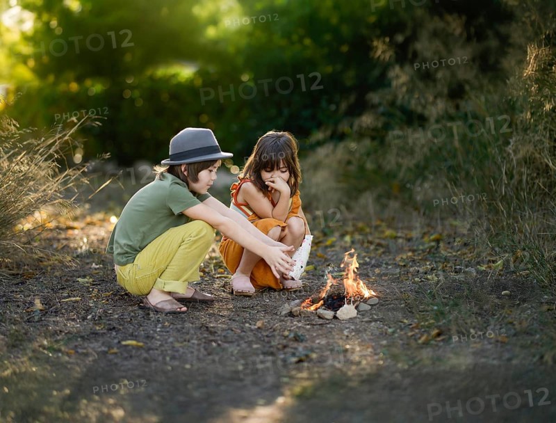 Two children sit by a small campfire in a woodland area. They are enjoying the outdoors, surrounded by nature. The setting sun creates a warm and peaceful atmosphere