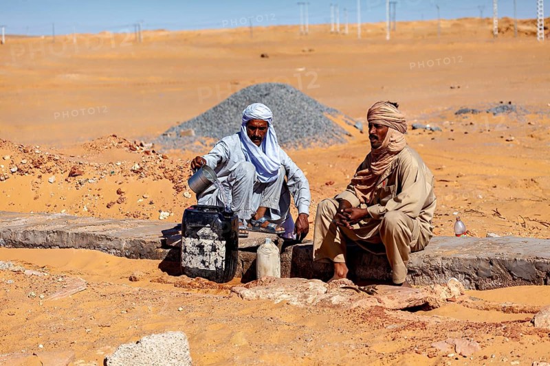 Two men in traditional dress sit in a sandy desert landscape, Two men draw water in the Sahara in Algeria