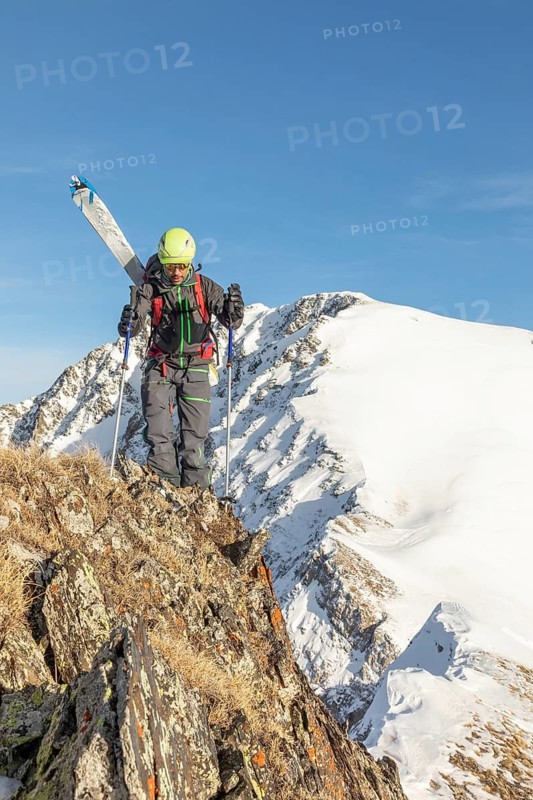 Skier in vibrant gear traverses rugged, snow-capped mountain terrain under a clear blue sky. The remote and challenging location highlights the thrill of ski touring