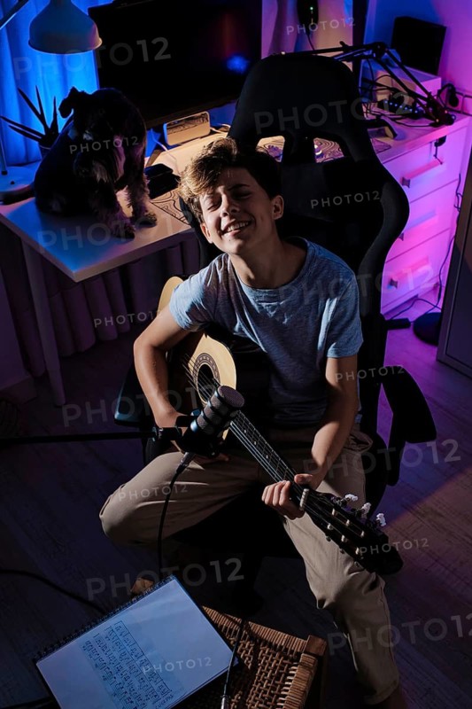 A young guitarist smiles while playing acoustic guitar in a cozy, vibrantly lit home studio with a microphone and a dog on the desk, creating an inviting musical atmosphere