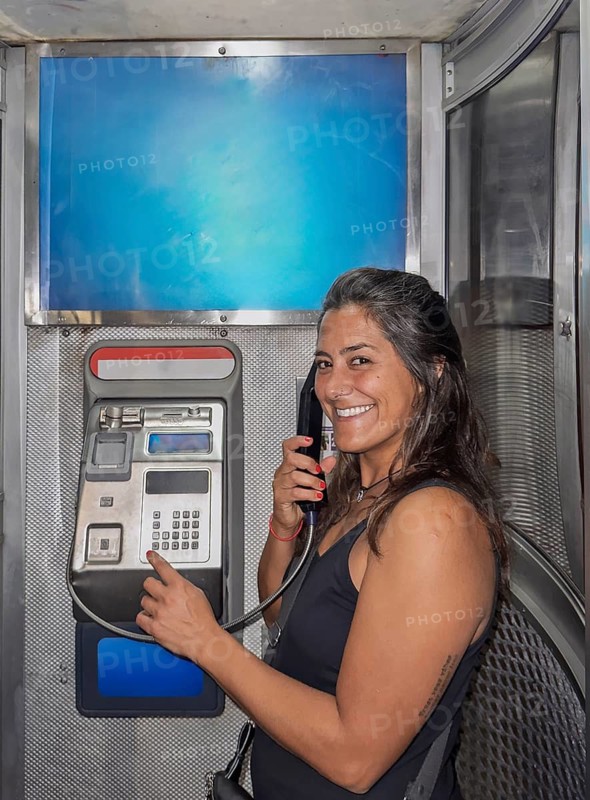 A smiling woman is using a public payphone inside a metallic booth located in Melbourne, Australia. The image captures a moment of nostalgia in modern urban life