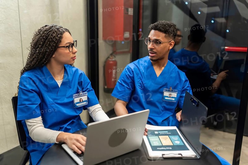 Medical staff in blue scrubs discussing patient information, collaborating on a laptop and reviewing documents in a contemporary hospital or clinic setting